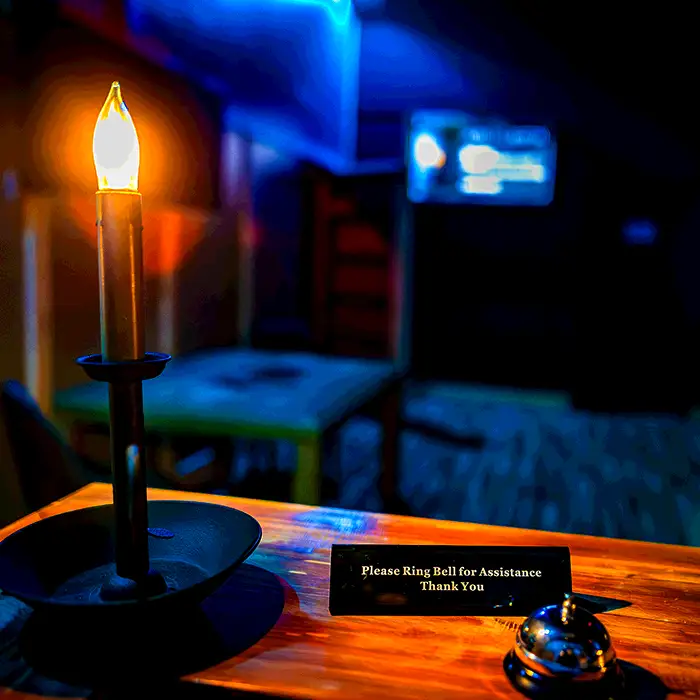 Candle on a wooden desk with a service bell and a 'Please Ring Bell for Assistance' sign in a dim blue-lit room. | Escape Code | Branson, Missouri Escape Room