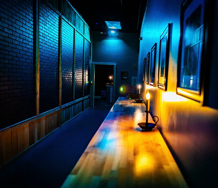 Dim hallway with blue neon lighting, a long wooden counter with a lamp, brick wall on the left, and framed pictures on the right. A doorway glows at the far end. | Escape Code | Branson, Missouri Escape Room